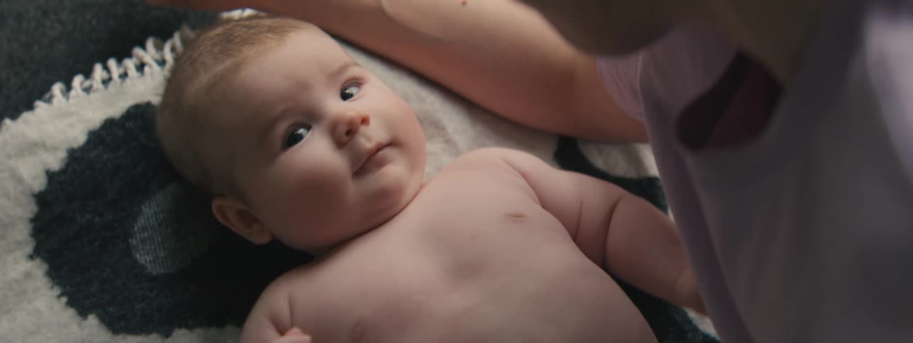 An infant lies on a soft surface with an adult gently talking to him