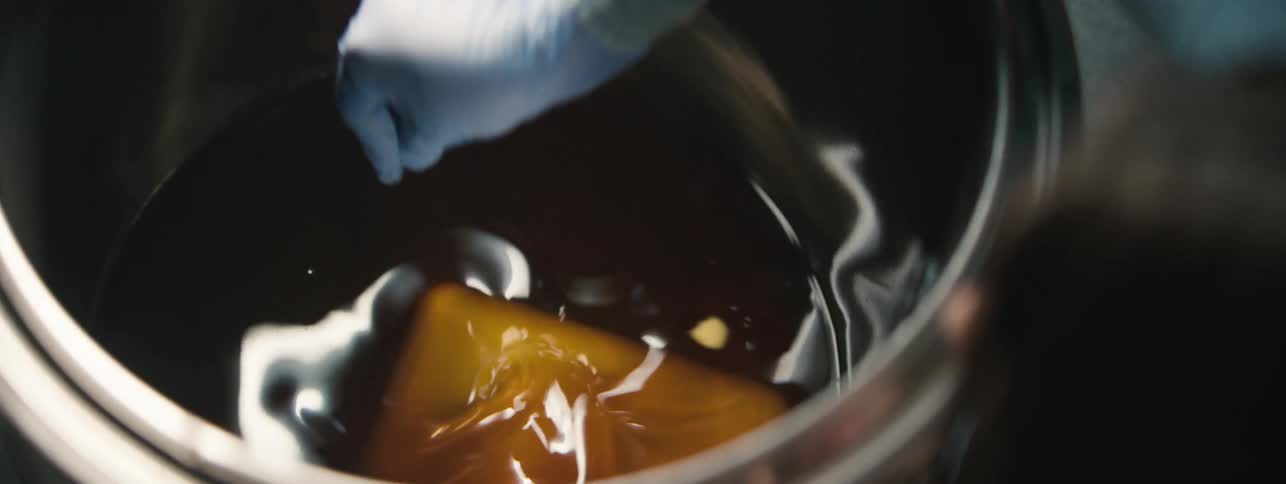 A gloved hand submerges a yellow block into an amber liquid inside a metal container