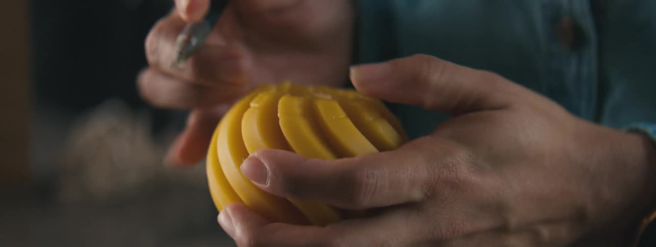 A persons hands using an iron tool to scrape an orange object into a carving shape