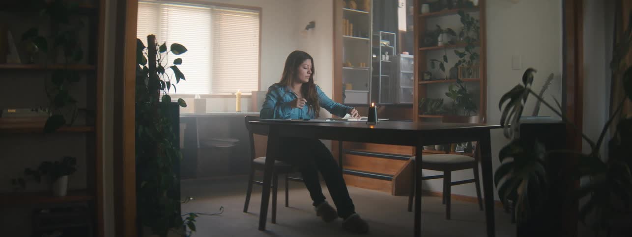 Awoman in a denim shirt working on a drawing at a desk in a warmly lit room