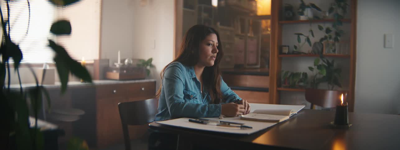 A closeup of a woman in a denim shirt working on a drawing at a desk in a warmly lit room