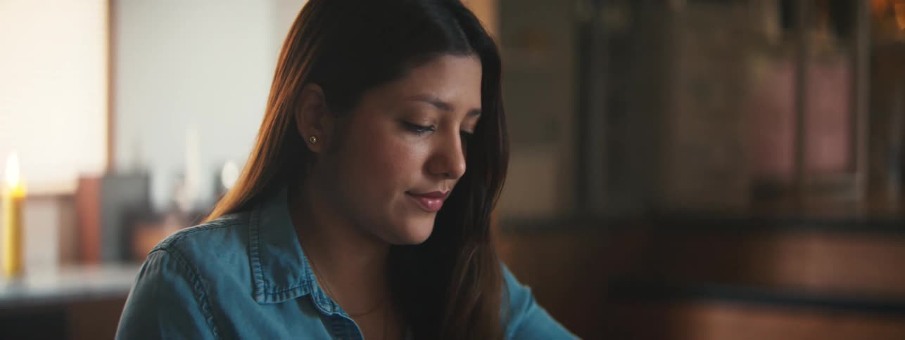 A closeup of a woman in a denim shirt working on a drawing at a desk in a warmly lit room
