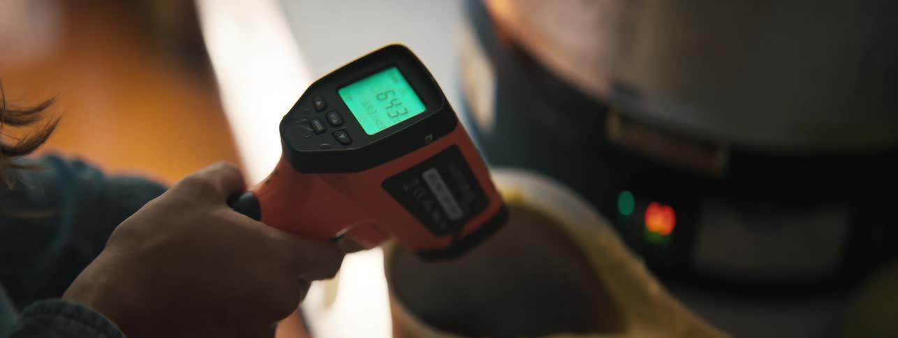 A closeup of a hand holding an infrared thermometer aimed at a jar filled with liquid