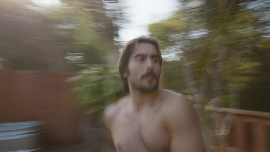 A strong shirtless muscular young man stretches in the backyard in the morning