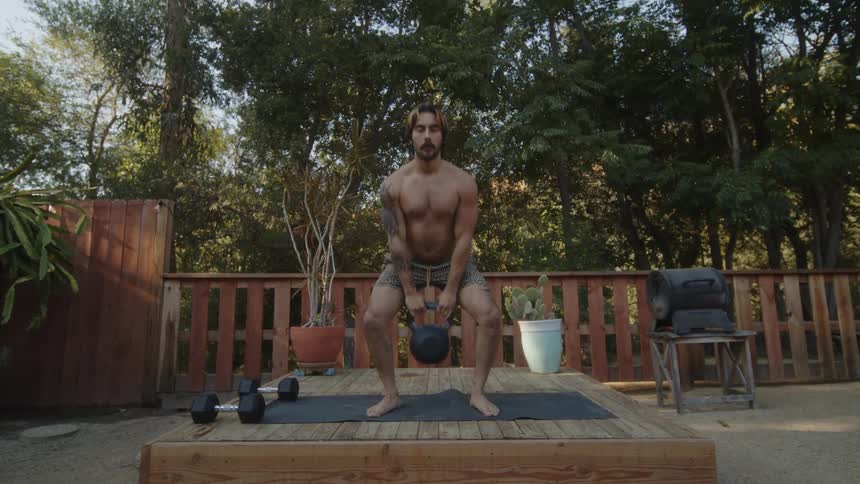 A man in shorts lifts a kettlebell while standing on a wooden platform on a backyard deck