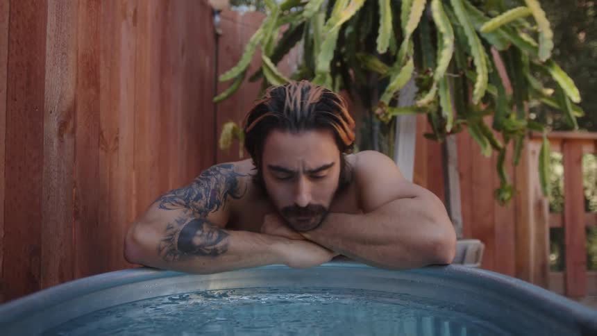 A man kneels next to an ice bath dipping his fingers in and running them through his hair then watching the ice melt in the water