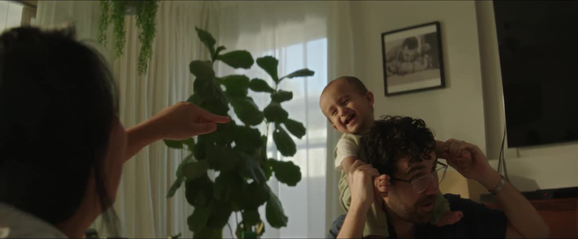 Mother gestures as father holds their baby on his shoulders while they sit at home