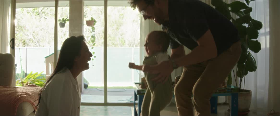 A father bounces his toddler son across a living room to his laughing mother