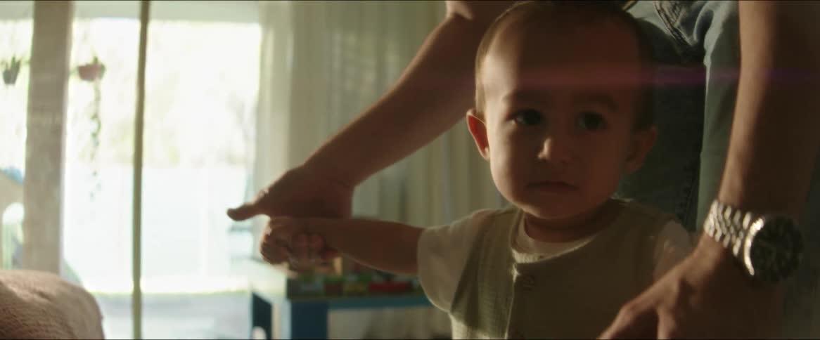 A father holds his toddler by the hands and moves him around in a living room