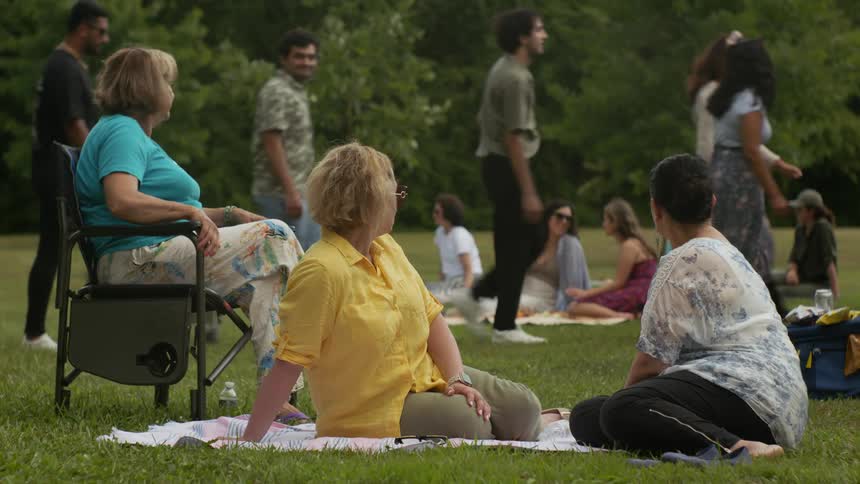Three women sitting in a park watch a group of people walk by