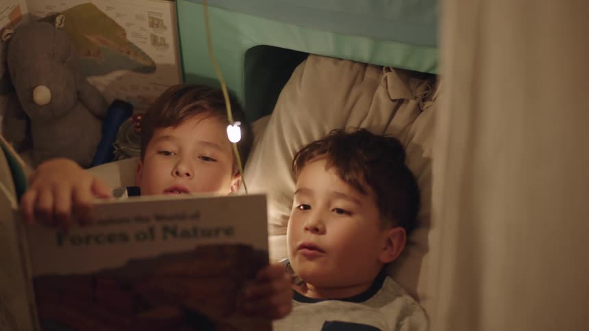 Two brothers lie in a bed with one holding a book and his brother reading out loud from it