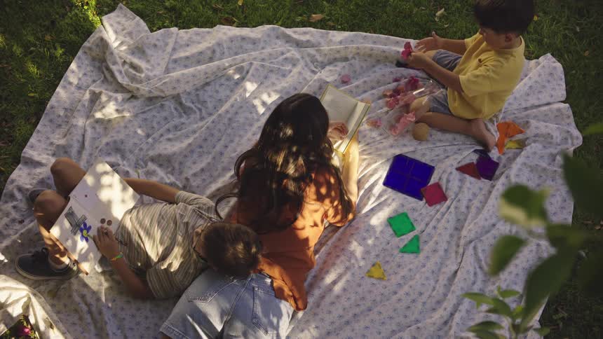 Mother lying on a blanket with her two children with one of them reading a book and the other playing