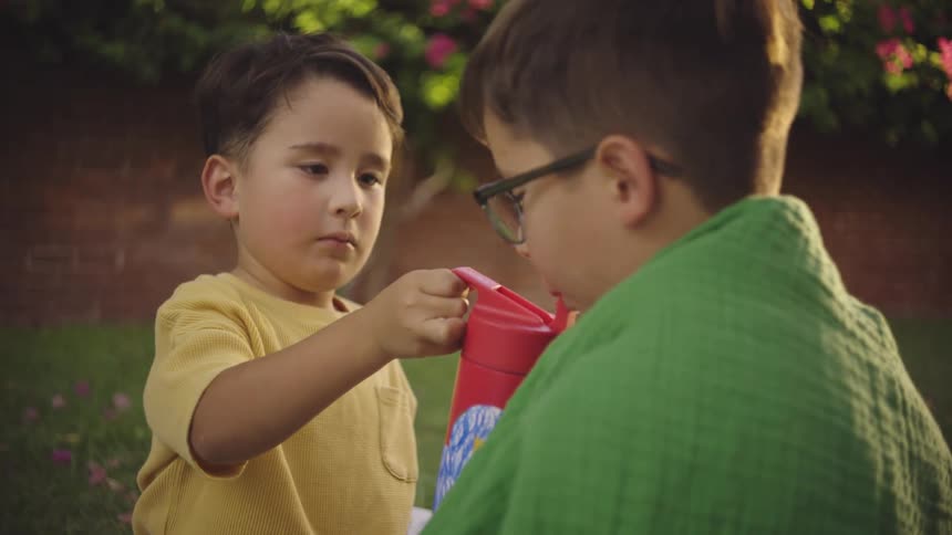 Two boys sitting in the backyard with one of them offering a drink in a red bottle to the other