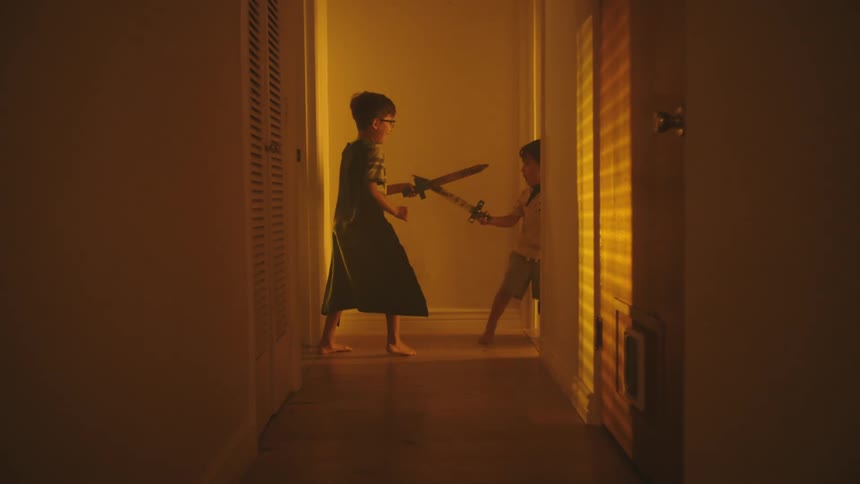 Two young boys wearing capes and holding toy swords are having a playful sword fight in a hallway