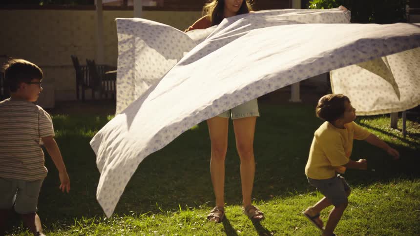A mother plays with a sheet and her two sons outdoors on a summer day