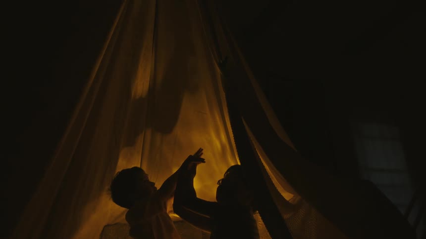 Two little boys in a sheet hut in a dark room playing shadow play with their hands