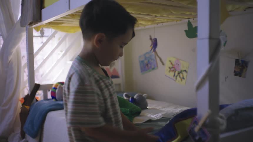 A young boy carries a green blanket to his bed and grabs a toy from underneath his pillow
