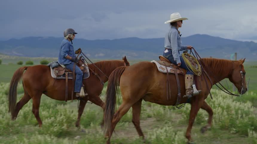 Two women on horseback trotting along a grassy field