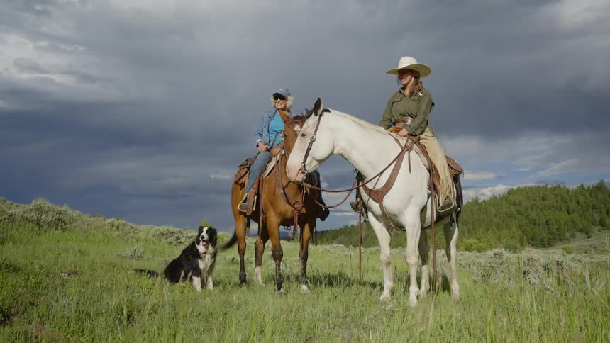 Two women on horseback stopped on a grassy plain with a dog sitting beside them as storm clouds gather behind them