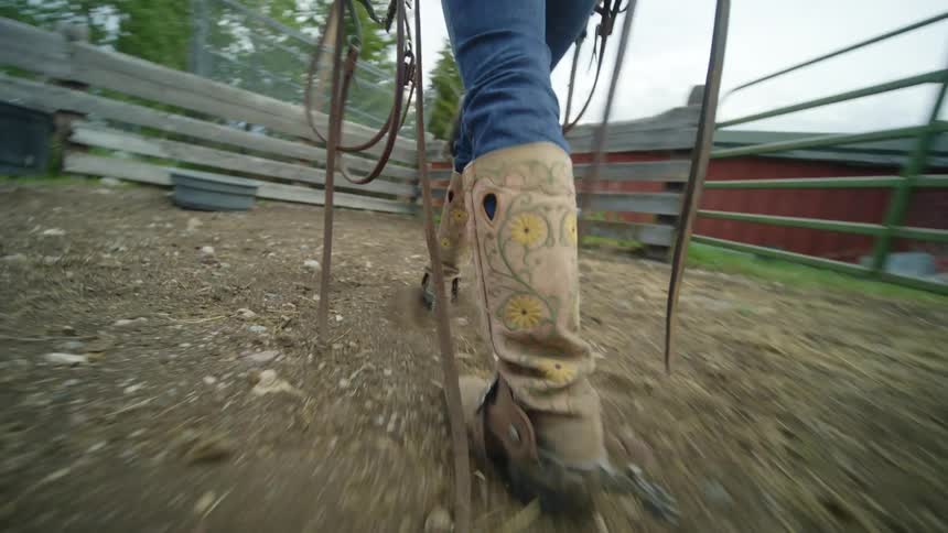 Someone wearing cowboy boots and carrying leather straps steps onto a corral and heads to a horse