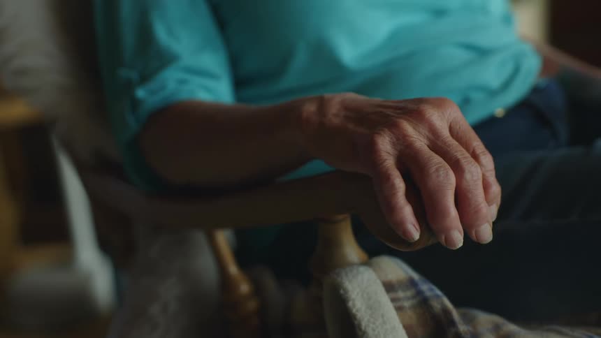 An elderly hand grasps the arm of the rocking chair on which it rests