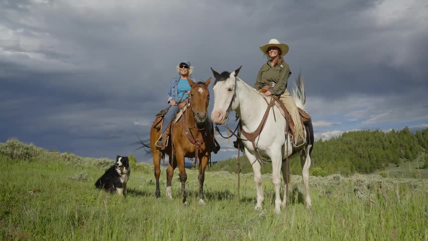 Two female ranchers on horses with a dog smile as they look into the distance outdoors