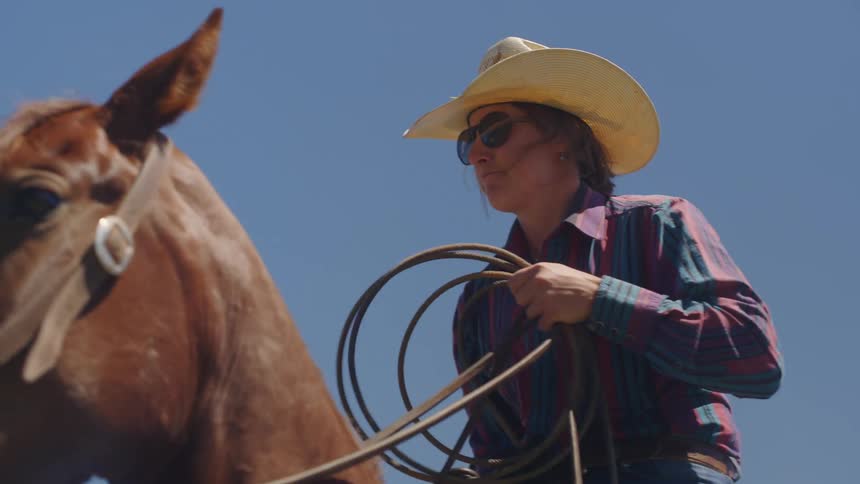 A woman wearing a cowboy hat and a plaid shirt holding a lasso while mounted on a brown horse