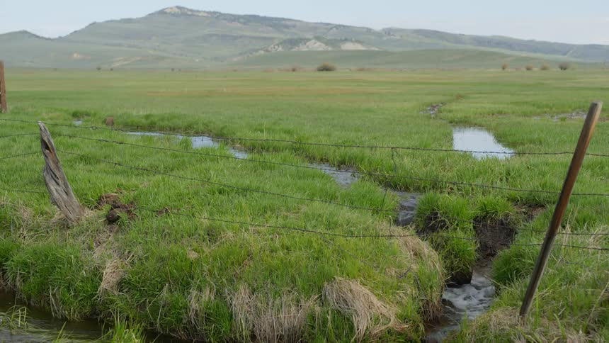 A small stream flows under a barbed wire fence on a plain