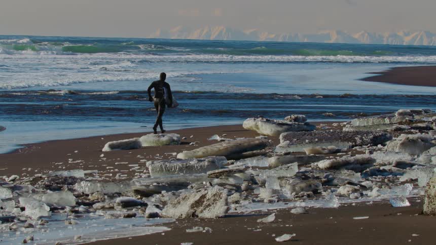 Surfer with surfboard running on sand with ice at seaside