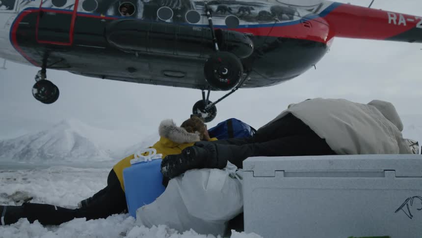 Three people lying in the snow and there is a white and red helicopter taking off