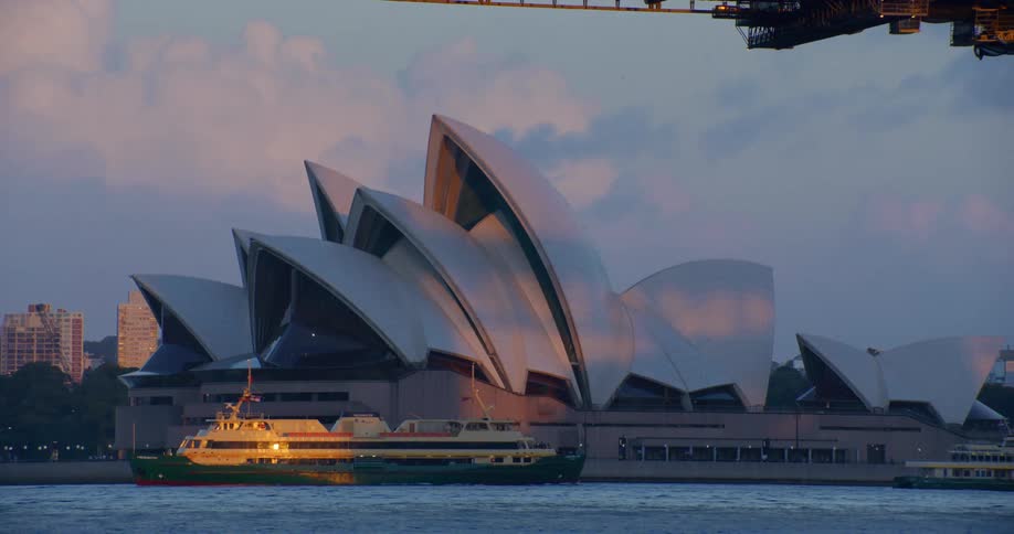A ferry passing in front of the opera house on the water at sunset