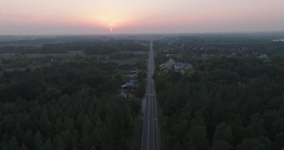 Aerial panorama of a long highway surrounded by vegetation and scattered houses on a day with a reddish sky and the sun on the horizon