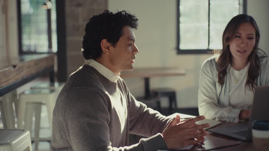 A man and a woman talk and smile at a cafeteria table with a laptop notepads and disposable coffee cups in front of them