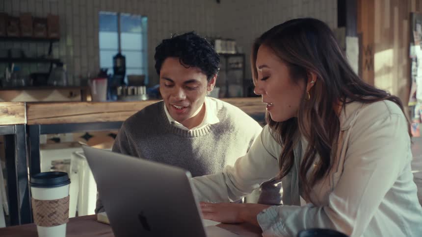 Man and woman sitting together at a table talking and writing with an open laptop