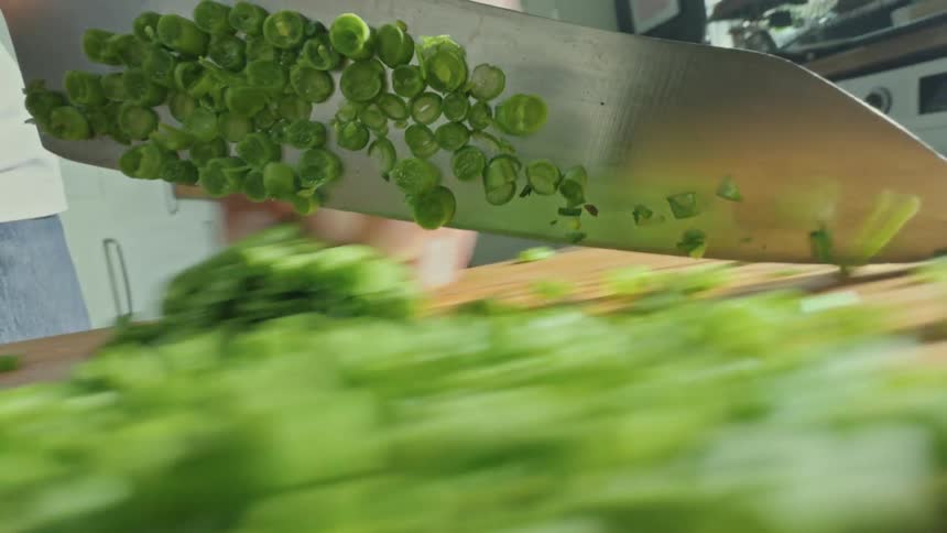 Point of view of a knife cutting green vegetable on top of a wooden cutting board