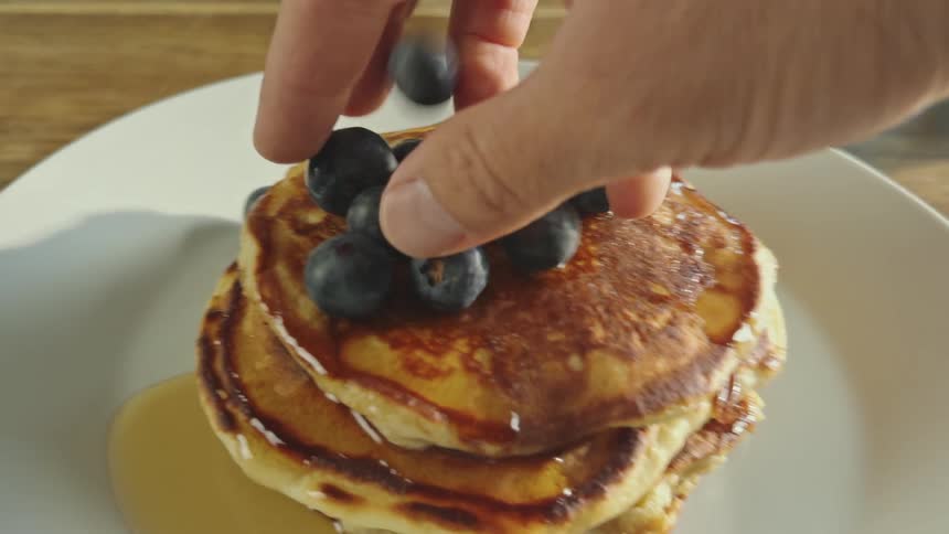 Close up point of view of a hand picking up blueberries and placing them on top of a plate of pancakes