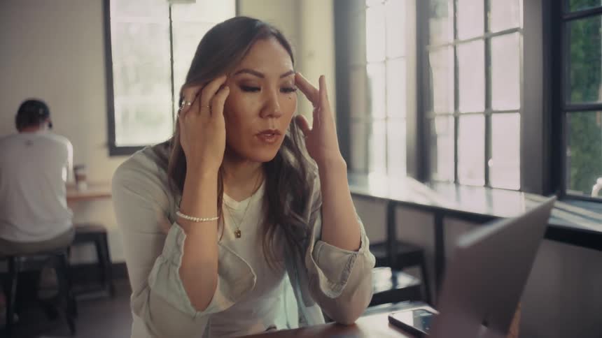 A woman sits at a desk near large windows massaging her temples to relax