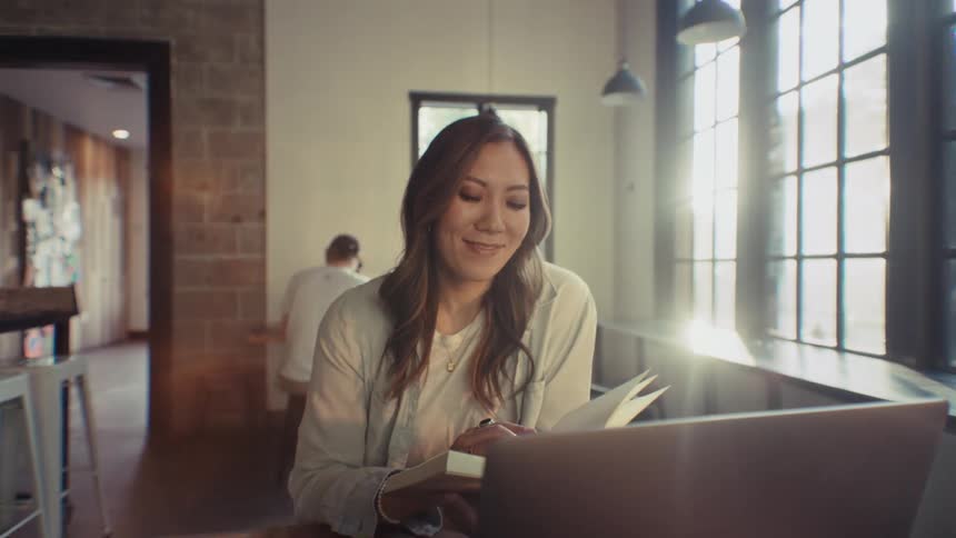 A woman looking at a book while sitting in front of a laptop and then she looks forward