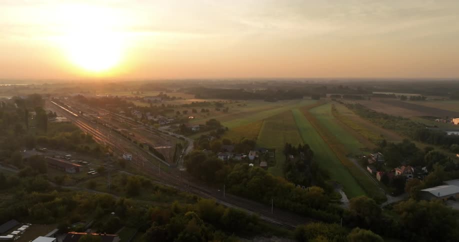 Aerial view of a train station with tracks surrounded by green fields and the sun on the horizon