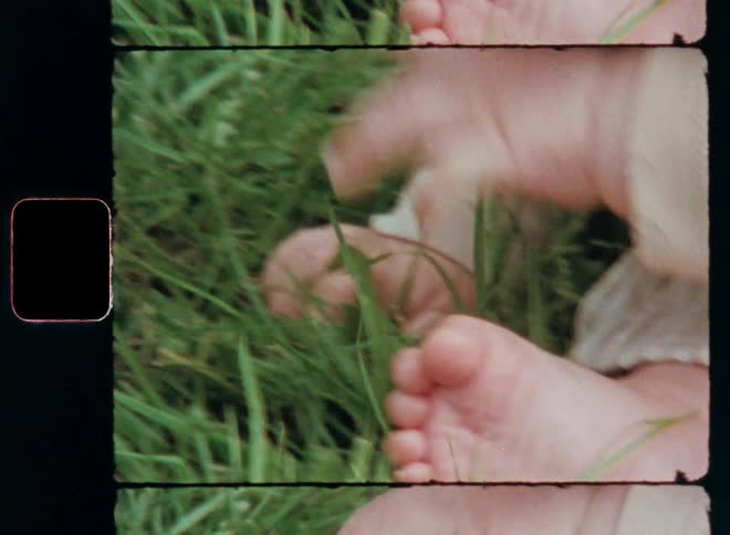 A cute baby is sitting on the ground touching the green grass with its bare feet and hands feeling the connection to nature expressing curiosity and great interest on vintage Super-8 film footage