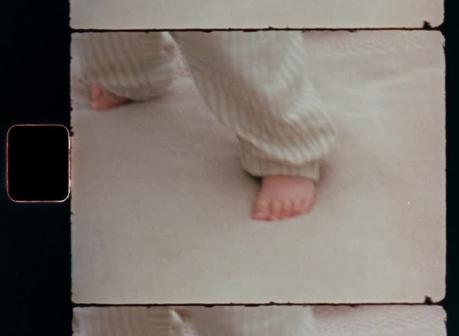 A mother or father is helping a baby girl stand on shaky or wobbly legs while holding or supporting the young child that is learning how to walk and making her first steps caught on a vintage home movie Super 8 film