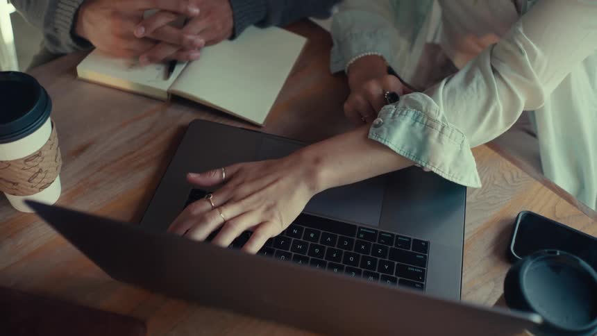 A womans hand types on a laptop while a mans hand rests on a notebook