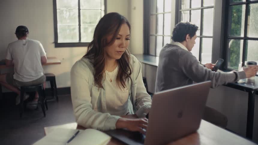 A woman with long hair typing on a laptop over a desk near large windows