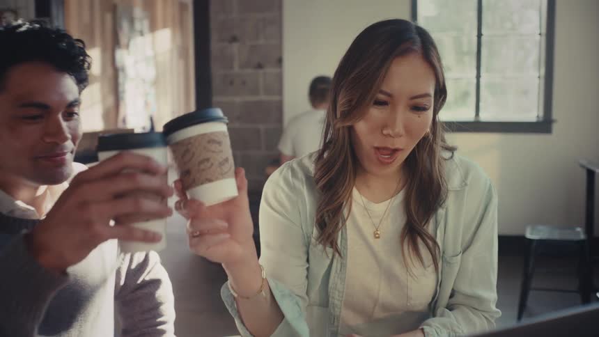 Cheerful man and woman sitting toasting with disposable coffee cup while sitting in front of laptop