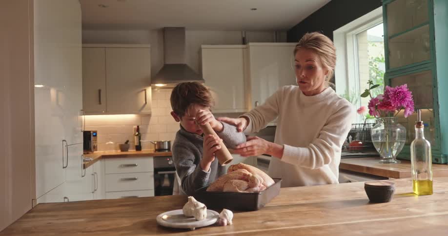 A mother keeps close tabs on her son as he tries to grind herbs onto a chicken in a pan