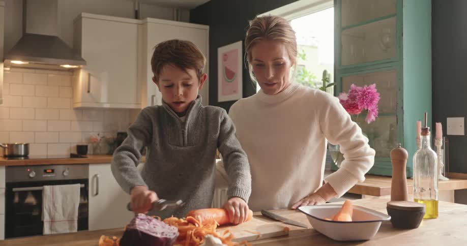 Young boy peeling carrots at the kitchen island as his mother watches