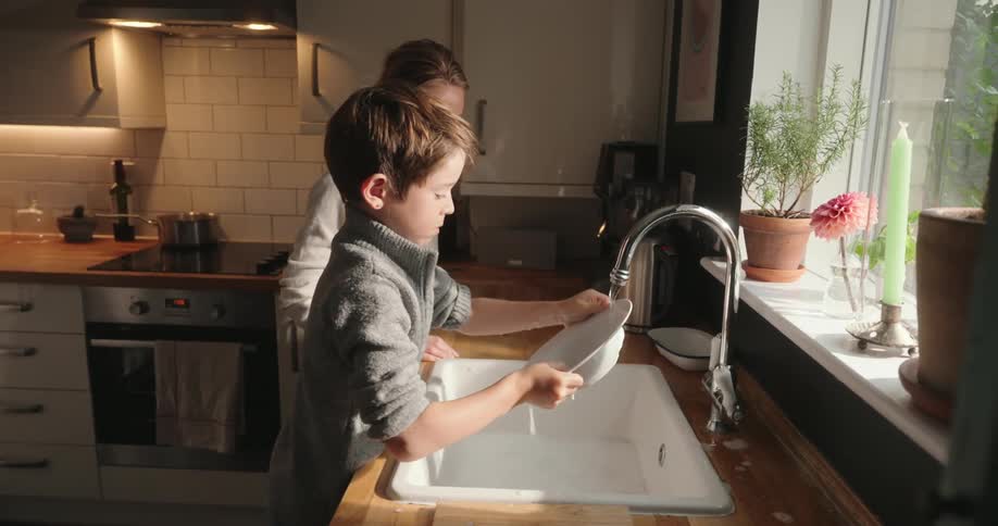 A mother smiles as she watches her male son wash a dish under the kitchen sink faucet