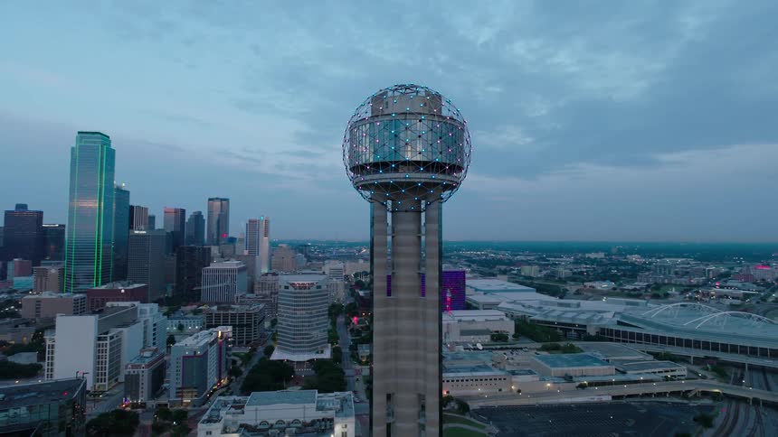 Cityscape at dusk with a prominent spherical structure atop a tower in the foreground and illuminated buildings in the background
