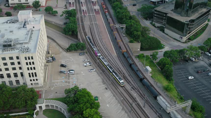 Aerial of a passenger train and a freight train passing between buildings during the daytime