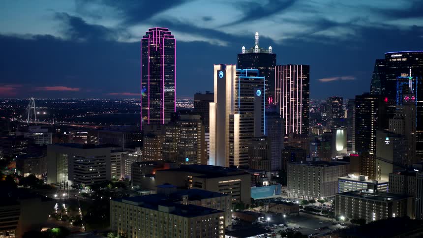 The center of a metropolitan city with tall buildings at night and there is a large building with pink lights on its exterior